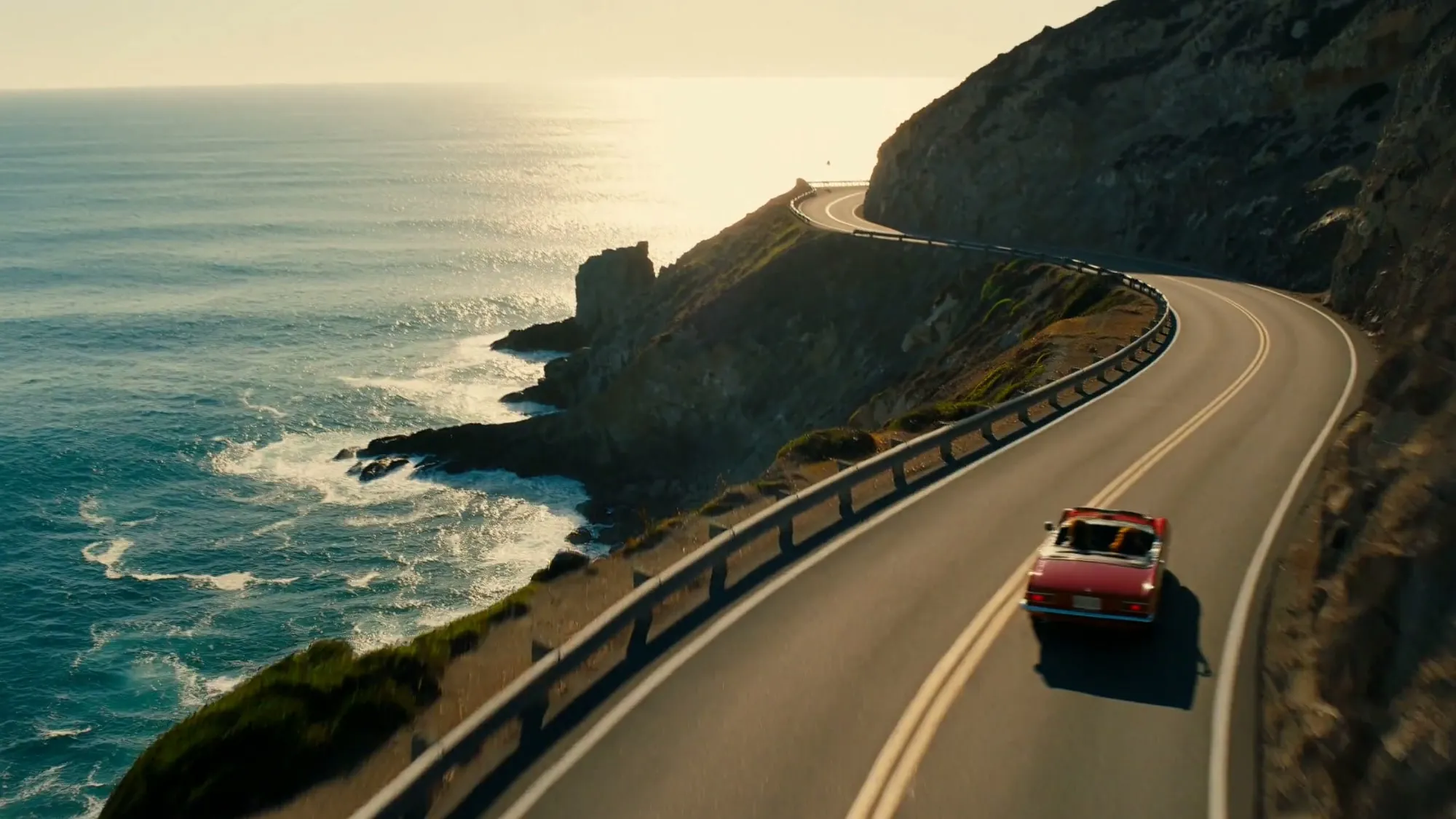 Un cabriolet rouge roule sur une route en lacets au-dessus d'une mer turquoise au coucher du soleil, vue aérienne
