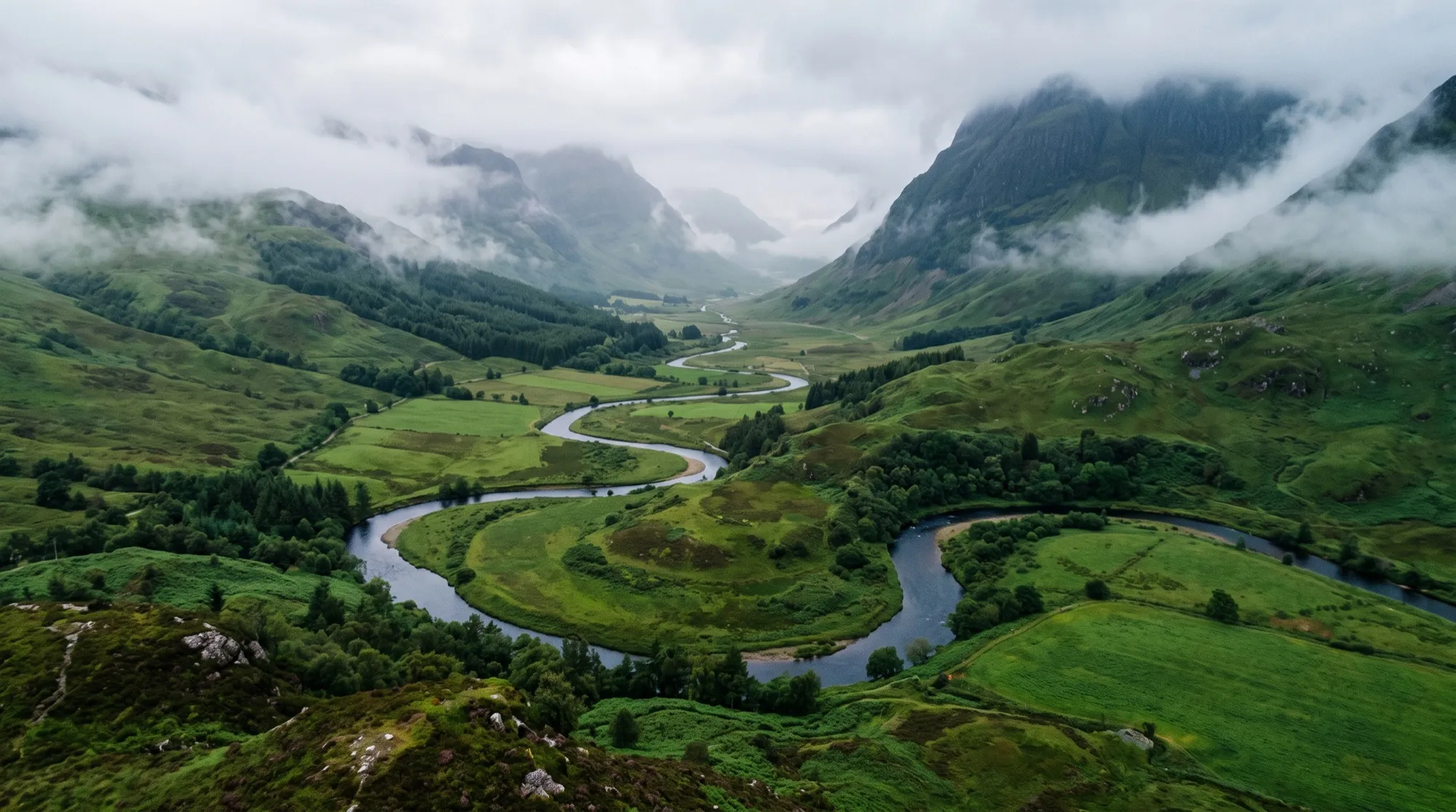 Vue aérienne d'une vallée verte brumeuse avec une rivière sinueuse et des nuages sur les pentes