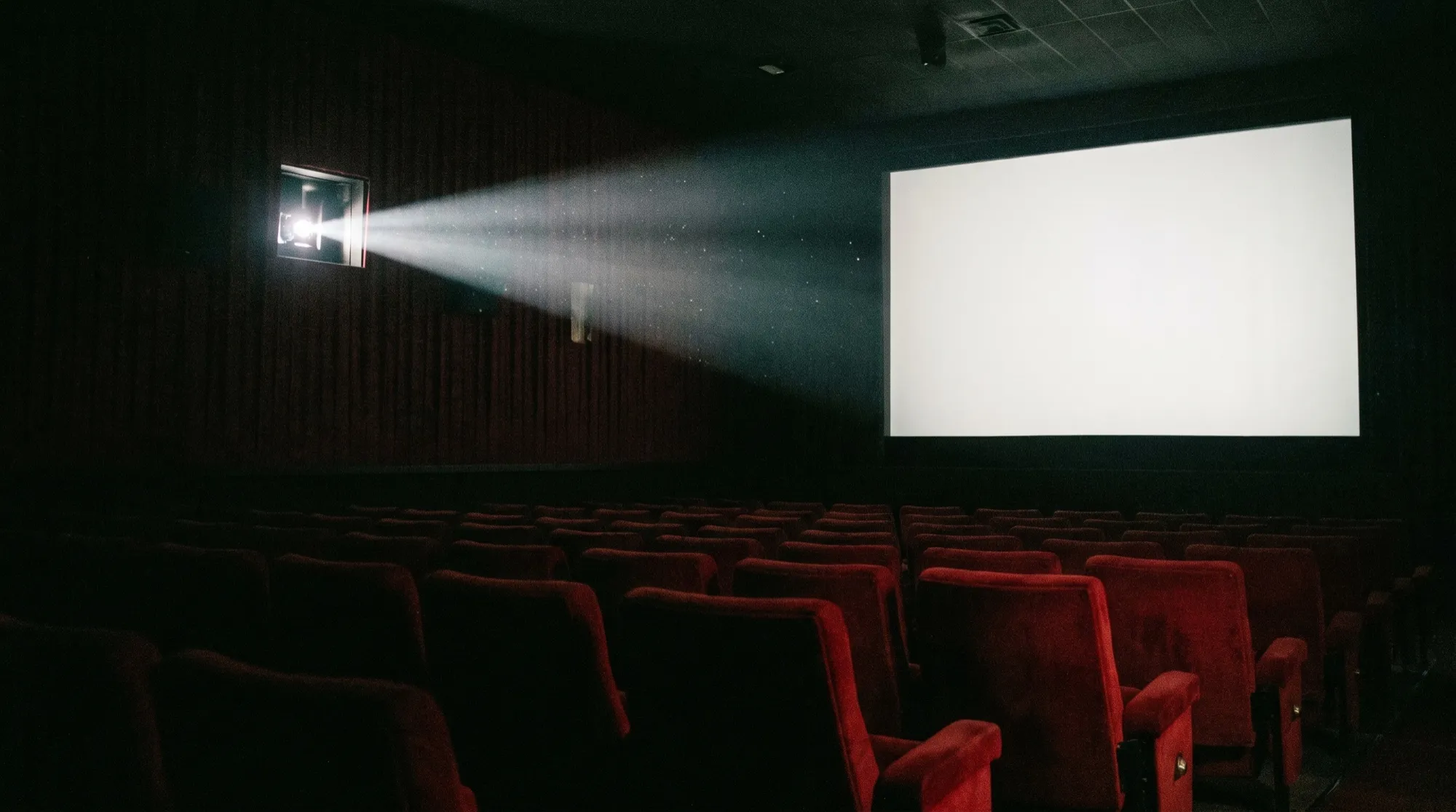 Une salle de cinéma vide et sombre : le faisceau du projecteur frappe un écran blanc, des rangées de fauteuils rouges