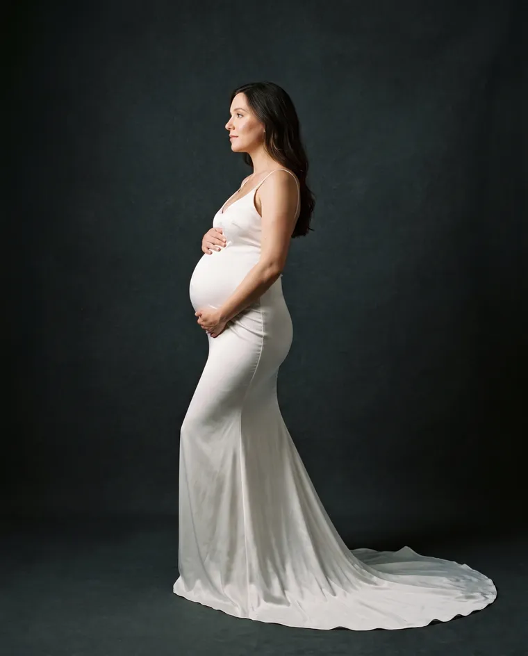Une femme enceinte en robe blanche ajustée à traîne, de profil sur fond graphite sombre, les mains sur le ventre