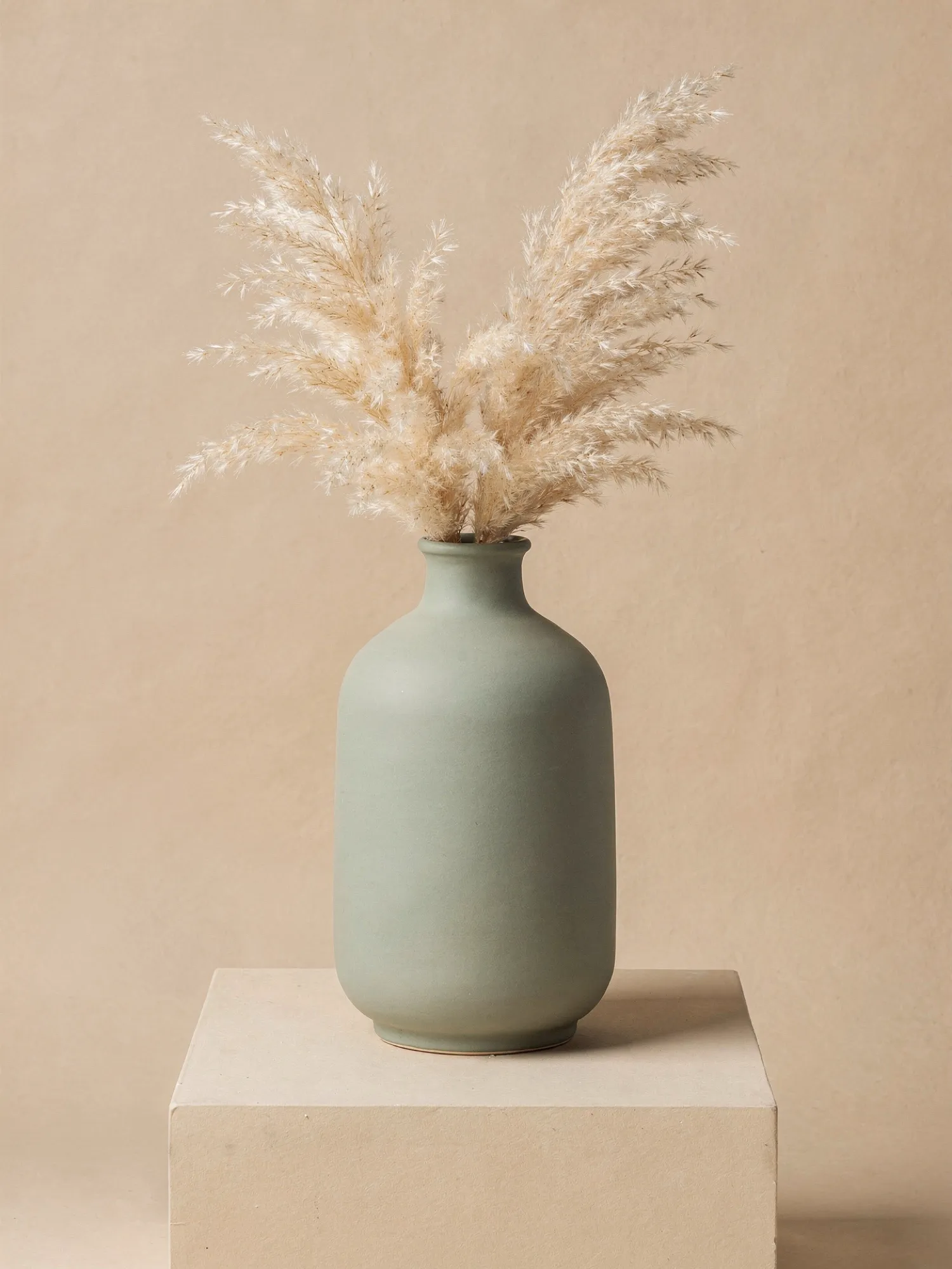 Un vase mat vert sauge avec de l'herbe de la pampa séchée sur un socle beige